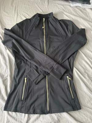 Svart träningsjacka från Lululemon - Svart figursydd träningsjacka från Lululemon med hel dragkedja i guld, två fickor med dragkedja och hög krage. Tillverkad i stretchigt funktionsmaterial som andas. Perfekt för gymmet eller löprundan.