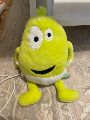 Babblarna Babba gosedjur - Rolig och mjuk gul plyschfigur med stora svarta och vita ögon, ett brett leende och vita detaljer. Figuren har armar, ben och en liten tofs på huvudet. Perfekt som prydnad eller kramkompis.