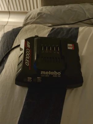Metabo batteriladdare ASC 55 - Svart batteriladdare från Metabo, modell ASC 55, för 12V-36V batterier. Laddaren har märkningen 'AIR COOLED' och CAS-system. Tillverkad i plast med tydliga logotyper och indikatorer på framsidan. Perfekt för laddning av verktygsbatterier.