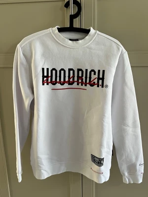 Vit crewneck från Hoodrich - Vit sweatshirt från Hoodrich med svart och rött tryck på bröstet och ryggen. Crewneck-modell med långa ärmar och ribbade muddar. Snygga detaljer med broderad text och patch nertill. Perfekt för en streetwear-look.