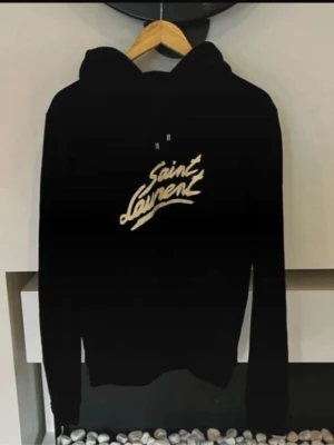 Svart Saint Laurent hoodie - Svart hoodie från Saint Laurent med vit logga framtill i snyggt skriven stil. Klassisk modell med huva och dragsnören samt en stor magficka. Perfekt för dig som gillar streetwear och vill ha något trendigt och bekvämt.
