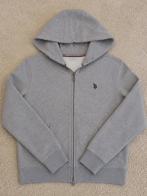 Grå zip-hoodie från U.S. Polo Assn. - Snygg grå hoodie med dragkedja från U.S. Polo Assn. Klassisk design med huva, två fickor fram och broderad logga på bröstet. Mjuk bomullsblandning och ribbade muddar för extra komfort. Perfekt för en chill och stilren look.