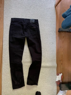 Svarta jeans från nudie (grim Tim ) - Säljer ett par svarta jeans från nudie med klassisk femficksdesign och raka ben. Jeansen har diskreta detaljer och logga på bakfickan. Materialet är stretchigt denim som sitter snyggt och bekvämt.