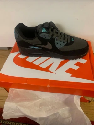 Nike Air Max 90 svart/turkos sneaker - Nike Air Max 90 sneakers i svart och mörkgrått med turkosa detaljer. Klassisk siluett med synlig Air Max-dämpning i sulan och robust yttersula. Ovandel i mesh och syntetmaterial, snörning framtill och ikonisk Swoosh-logga på sidan.