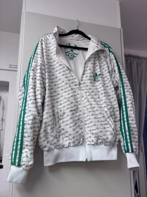 Vit Adidas vindjacka med gröna ränder - Säljer en vit vindjacka från Adidas med gröna ränder längs ärmarna och all-over logomönster. Jackan har dragkedja framtill, hög krage och ribbade muddar. Perfekt för dig som gillar sportig och retro stil. Behöver en tvätt