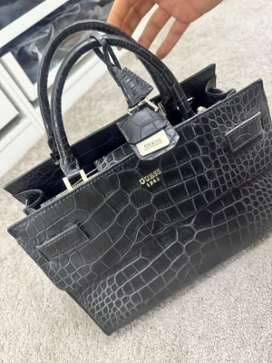 Svart croco handväska från Guess 350kr - Snygg svart handväska från Guess i croco-mönstrat konstläder. Väskan har två rejäla handtag, silverfärgade detaljer och flera fack med dragkedja. Perfekt för dig som gillar en stilren och trendig look med lyxig känsla.