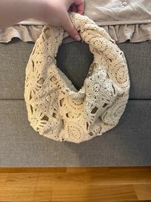 Beige virkad handväska - Superfin beige handväska i virkat material med rundad form och bred axelrem. Väskan har ett unikt mönster med öppna partier och känns både bohemisk och trendig. Perfekt accessoar för att lyfta din outfit.