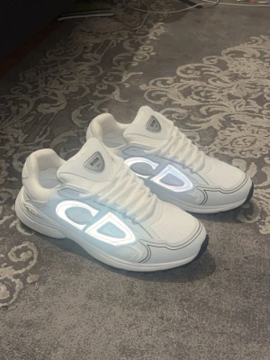 Vita CD B30 Skor - Snygga vita sneakers med stora reflekterande CD-detaljer på sidorna. Perfekta för dig som vill sticka ut med exklusiv streetstyle.