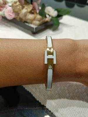Vitt armband med gulddetaljer - Snyggt armband i vitt med guldfärgade detaljer och en stor H-formad dekoration framtill. Rostfritt stål och nickelfritt. 6 cm diameter. Oanvänd 