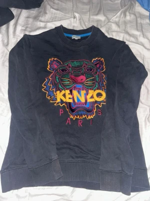 Svart Kenzo sweatshirt med tiger - Cool svart sweatshirt från Kenzo med ikonisk broderad tiger i flera färger och texten 'KENZO PARIS' framtill. Tröjan har rund halsringning och långa ärmar. Perfekt statement-plagg för dig som gillar streetwear och färgglada detaljer. Tröjan är storlek XS men passar som S.
