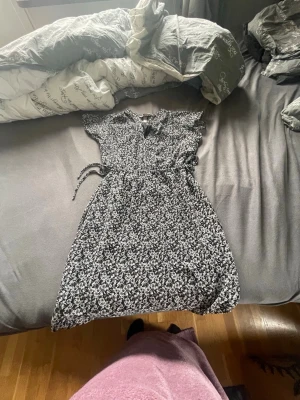 Svartvit blommig omlottklänning - Snygg svartvit omlottklänning med blommigt mönster och knyt i midjan. Klänningen har korta ärmar och en lätt v-ringning. Perfekt för dig som gillar en feminin och avslappnad stil.