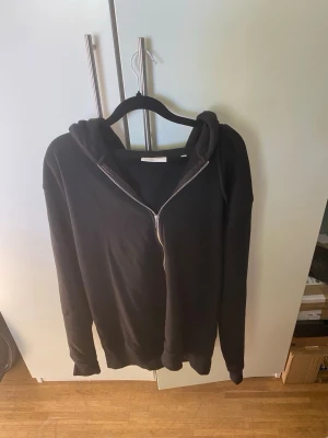 Svart hoodie med dragkedja Lindbergh XL - Svart hoodie från Lindbergh i relaxed fit med dragkedja framtill och huva. Perfekt för chill dagar och enkel att matcha med jeans eller joggers. Materialet känns mjukt och bekvämt, och modellen har långa ärmar och klassisk design.
