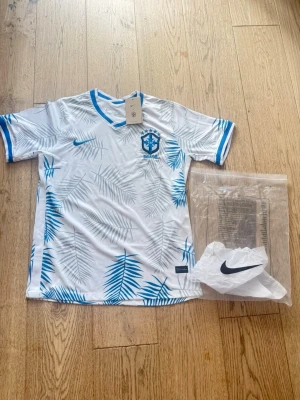Vit Nike Brasilien fotbollströja - Snygg vit fotbollströja från Nike med blå palmbladsmönster och blå detaljer vid krage och ärmslut. Brasilien-emblem på bröstet och klassisk Nike-logga. Perfekt för träning eller match. Tillverkad i lätt och ventilerande material. Hör av dig eventuella funderingar!