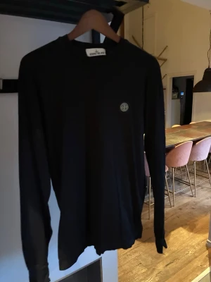 Svart långärmad tröja Stone Island - Svart långärmad tröja från Stone Island med rund hals och klassisk logotyp på bröstet. Tröjan har en stilren och enkel design, perfekt för dig som gillar streetwear. Materialet är mjukt och bekvämt, passar till många olika outfits.