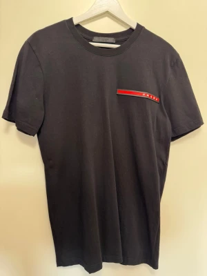 Svart t-shirt  - Snygg svart t-shirt  rund hals och korta ärmar. På bröstet finns en diskret röd och vit logodetalj som ger en exklusiv känsla. Tillverkad i mjuk bomull för maximal komfort och stilren look.