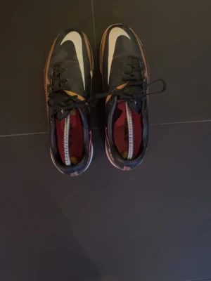 Nike svart/guld träningsskor  - Säljer ett par snygga Nike fotbollsskor i svart med guldiga och vita detaljer samt rosa logga på sidan. Skorna har låg profil, snörning och är tillverkade i syntetmaterial. Perfekta för dig som vill sticka ut på planen.
