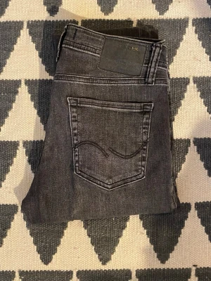 Svarta slim fit jeans från Jack n Jones  - Säljer ett par svarta slim fit jeans från jacknjones Jeans med slitningar och tvättad look. Jeansen har klassisk femficksmodell, normalhög midja och smala ben. Materialet är mjukt denim med stretch för extra komfort.