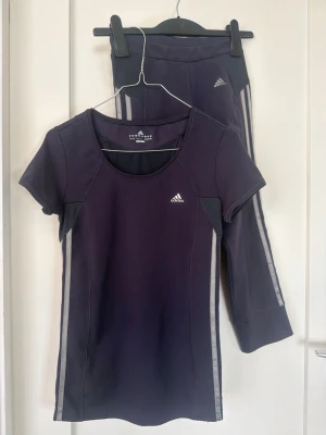 Mörklila träningsset från Adidas - Snyggt träningsset från Adidas i mörklila med silvriga ränder längs sidorna. Setet består av en kortärmad topp och matchande tights, båda med Adidas-logga på bröst och höft. Materialet är stretchigt och andas, perfekt för gym eller löpning.