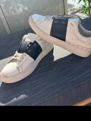 Valentino vita sneakers med svart detalj - Snygga vita sneakers från Valentino med bred svart läderdetalj över mitten och klassiska vita snören. Skorna har en stilren och modern design med rund tå och diskret logga på plösen. Perfekta för dig som gillar exklusiva märken och streetstyle.