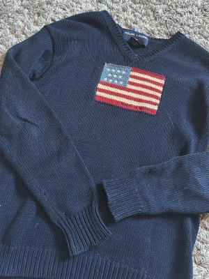 Mörkblå stickad tröja Ralph Lauren - Mörkblå stickad tröja från Ralph Lauren med V-ringning och en stor amerikansk flagga broderad på bröstet. Tröjan är gjord i 100% bomull och har ribbstickade muddar vid ärmslut och nederkant. Perfekt för dig som gillar klassisk preppy stil.