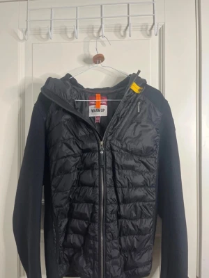 Svart pufferjacka från Parajumpers XS - Snygg svart pufferjacka från Parajumpers med huva och dragkedja framtill. Jackan har stickade ärmar och vadderad front, samt en gul detalj vid kragen. Perfekt för kalla dagar och har en sportig look.