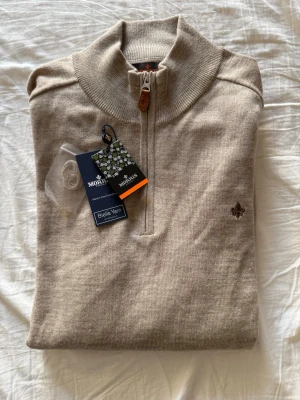 Beige halvzip-tröja från Morris - Snygg beige stickad tröja från Morris i 100% merinoull. Tröjan har halv dragkedja med läderdetalj, hög krage och en liten broderad logga på bröstet. Perfekt för lager-på-lager och stilren look. Aldrig använd. Är som en storlek L