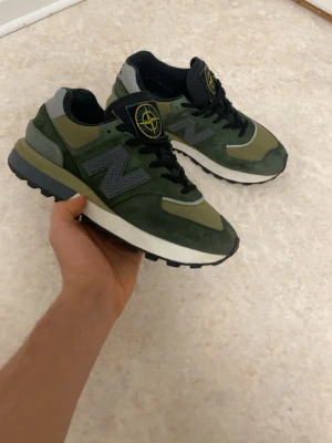 Gröna New Balance x Stone Island sneakers - Snygga gröna sneakers från New Balance i samarbete med Stone Island. Skorna har en mix av mocka och mesh, svarta och grå detaljer samt den ikoniska Stone Island-loggan på plösen. Perfekta för dig som vill sticka ut med stilrena och unika sneakers. Pris kan diskutera 