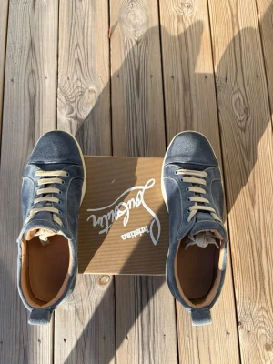 Ljusblå Louboutin sneakers med röd sula - Snygga sneakers från Christian Louboutin i ljusblå mocka med beigea skosnören och ikonisk röd sula. Låg modell med rund tå och diskreta sömmar. Perfekta för dig som vill sticka ut med exklusiva detaljer och klassisk design.