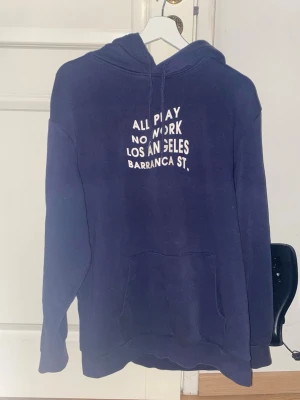 Mörkblå hoodie med tryck från Divided - Mörkblå hoodie med vit text på bröstet. Klassisk modell med huva och känguruficka framtill. Mjuk bomullskänsla och avslappnad passform, perfekt för en chill look. Storlek M