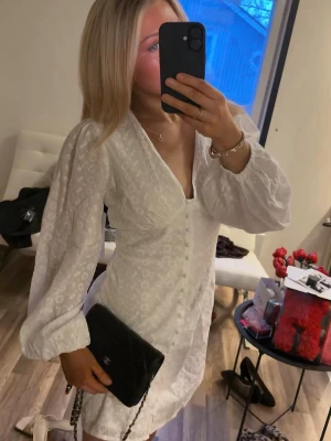 Vit kort klänning med broderad spets - Säljer helt ny vit kort klänning med v-ringning och broderad spets. Klänningen har puffiga långärmar och en slits framtill. Perfekt för sommarens alla tillfällen, och ger en fräsch och romantisk look.