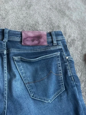 Blå jeans från Jacob Cohen - Snygga blå jeans från Jacob Cohen med klassisk femficksmodell och knappgylf. Jeansen har en mörkblå tvätt, kontrastsömmar och ett lila patch bak i midjan. Materialet är mjukt denim i bomull med stretch för extra komfort.