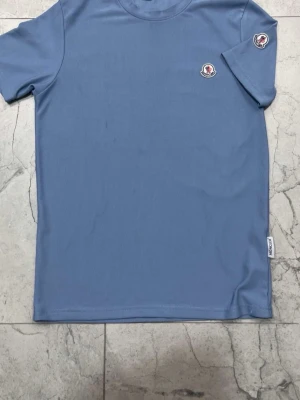 Blå Moncler t-shirt med logga - Snygg blå t-shirt från Moncler med rund hals och korta ärmar. T-shirten har två broderade Moncler-loggor, en på bröstet och en på ärmen. Tillverkad i mjuk bomull för en bekväm känsla. Perfekt för en stilren och avslappnad look.