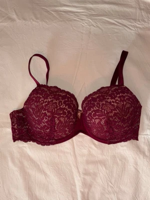 Vinröd spets-bh med vaddering - Oanvänd,Snygg vinröd bh med vadderade kupor och vacker spetsdetalj över hela framsidan. Justerbara axelband och hakknäppning bak,  tyvärr saknas en ögla bak sen bild 2 gjorde det när jag köpte den men det påverkar inte funktionen. Insidan är mjukt fodrad i ljusrosa tyg för extra komfort. Perfekt för dig som vill ha både stöd och stil.