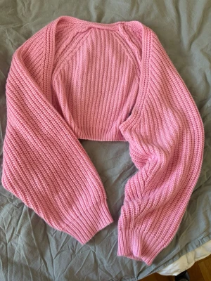 Rosa stickad bolero med vida ärmar - Säljer en söt rosa stickad bolero med ribbstickat mönster och långa, vida ärmar. Perfekt att slänga över en topp för en extra touch. Materialet känns mjukt och mysigt, och färgen är riktigt poppig.