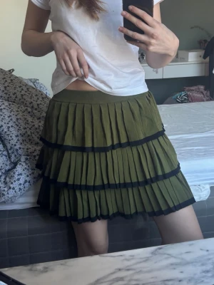 Olivgrön plisserad volangkjol - Superfin olivgrön kortkjol med flera lager volanger och svarta kanter. Kjolen är plisserad och har en bred, bekväm midjeresår. Från Kate Moss i nyskick 