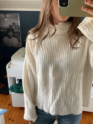 Vit ribbstickad polotröja - Säljer en vit ribbstickad polotröja med hög krage och långa ärmar. Tröjan har en lös passform och är gjord i ett mjukt stickat material som känns skönt mot huden. Perfekt för att skapa en avslappnad och trendig look.