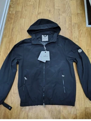 Svart vindjacka från Moncler - Snygg svart vindjacka från Moncler med huva och dragkedja framtill. Jackan har två framfickor med dragkedja och Moncler-logga på vänster ärm. Tillverkad i ett lätt, vattenavvisande material som passar perfekt för blåsiga dagar.