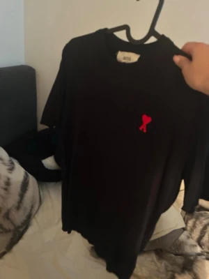 Svart t-shirt från AMI Paris - Svart stickad t-shirt från AMI Paris med klassisk passform och korta ärmar. På bröstet sitter ett rött hjärta med bokstaven A, vilket är märkets ikoniska logga. Tillverkad i mjuk bomull för en skön känsla.