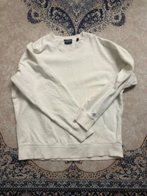 Vit sweatshirt från GANT - Vit sweatshirt från GANT i relaxed fit med rund halsringning och ribbade muddar. Klassisk och stilren design med liten logga på ärmen. Perfekt för en clean och avslappnad look.