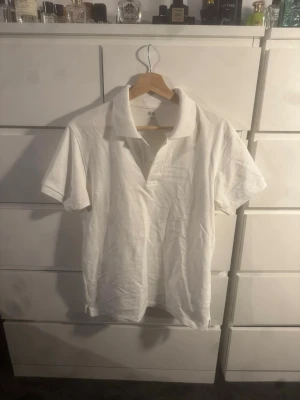 Vit pikétröja från Uniqlo - Klassisk vit pikétröja från Uniqlo med krage och korta ärmar. Tröjan har knappar vid halsen och en liten bröstficka. Perfekt till både jeans och shorts för en clean och enkel stil.