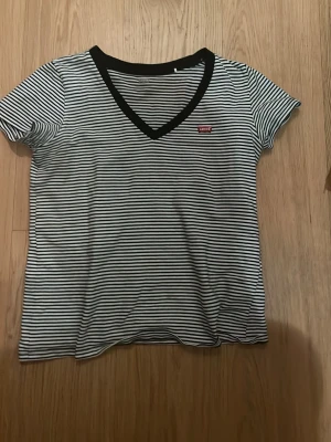 Randig v-ringad t-shirt från Levi's - Snygg t-shirt från Levi's med svartvita ränder och v-ringning med svart kant. Klassisk passform och liten röd Levi's-logga på bröstet. Perfekt till jeans eller shorts 