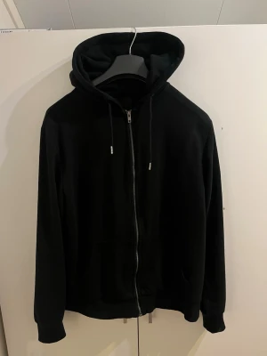Svart hoodie med dragkedja från 157 - Svart hoodie från 157 med dragkedja framtill, huva med dragsko och två fickor. Tillverkad i mjukt material som känns skönt mot huden. Perfekt för en avslappnad och stilren look.
