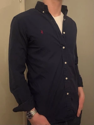 Ralph Lauren skjorta – S/M slim fit  - Säljer denna snygga blå Ralph Lauren skjorta i storlek S, Custom Fit vilket motsvarar ungefär M Slim Fit. Den är i bra skick förutom en liten missfärgning vid Ralph Lauren-loggan, då jag råkat stryka på den. Om du har några frågor tveka inte att höra av dig. Allt gott Deluxe Garderoben. 