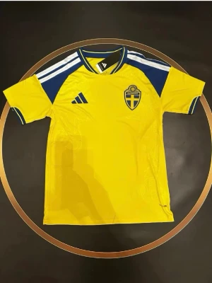 Sverige fotbollströja Adidas gul/blå - Gul fotbollströja från Adidas med blå detaljer och Sveriges landslagsemblem på bröstet. Tröjan har korta ärmar, rund hals och klassiska Adidas-ränder på axlarna. Perfekt för match eller träning. Materialet är lätt och andas.