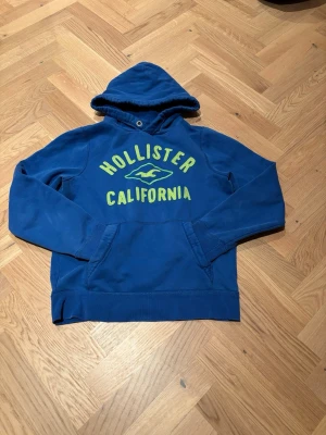 Blå Hollister hoodie med tryck - Blå hoodie från Hollister med broderat gult tryck 'Hollister California' och logga på bröstet. Klassisk känguruficka framtill och huva med dragsko. Perfekt för en avslappnad och cool stil. Riktigt snygg nu till våren med den blåa färgen!