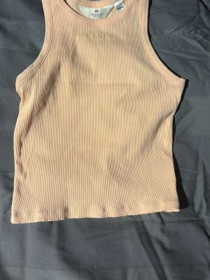 Rosa beige ribbad linnetopp från H&M Pantone - Snygg ribbad linnetopp från H&M i samarbete med Pantone. Toppen är rosa beige, ärmlös och har en hög halsringning. Perfekt för att styla med jeans eller kjol. Mjuk och stretchig bomullskvalitet.