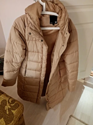 Beige lång dunjacka med huva - Snygg beige dunjacka med quiltat mönster och huva. Jackan är lång, har tryckknappar och dragkedja framtill samt två fickor. Perfekt för kalla dagar när du vill hålla dig varm och stilren.