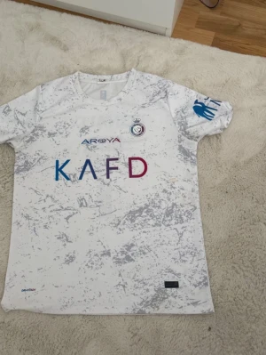 Al Nassr vit matchtröja - Snygg vit matchtröja från Al Nassr med grått marmormönster och klubbmärke på bröstet. Tryck i blått och vinrött framtill samt blått tryck på ärmen. Perfekt för dig som gillar fotboll och vill sticka ut på planen.