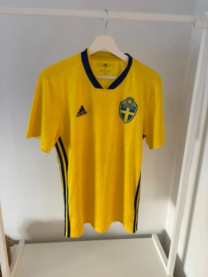 Svensk landslagströja - VM 2018 - Gul kortärmad fotbollströja från Adidas med Sveriges landslagsemblem på bröstet och blå detaljer vid krage och sidor. Tröjan har svart Adidas-logga och texten 'Sverige' i nacken. Perfekt för match eller träning. ÄKTA