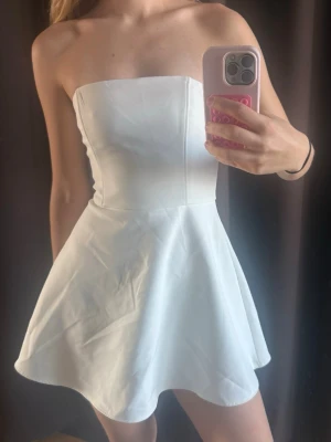 Vit axelbandslös kort klänning - Snygg vit axelbandslös klänning med figurnära överdel och utsvängd kjol. Perfekt för sommarens alla tillfällen. Tillverkad i ett mjukt material som ger en bekväm passform och stilren look.